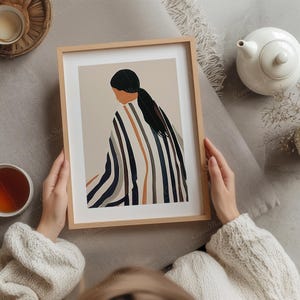May include: A framed art print featuring an abstract illustration of a person with a dark ponytail and a striped garment in shades of black, white, and brown. The artwork is held by a person wearing a white sweater, with tea and a teapot in the background.