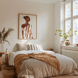 May include: A bedroom scene with a bed covered in a beige comforter and a brown throw blanket. A framed artwork of a person in a striped garment hangs above the bed. A small wooden stool and a vase with dried plants are also visible.