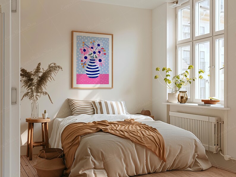 May include: A framed art print featuring a vase with blue and white stripes filled with pink and blue flowers. The artwork hangs above a bed with striped pillows and a brown blanket. The room has a neutral color palette with a window and a small wooden table.