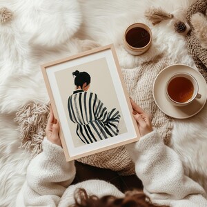 May include: A framed art print featuring a woman in a striped robe, viewed from the back, with a bun hairstyle. The artwork is held by a person wearing a white sweater, with a cup of tea and a decorative blanket in the background.