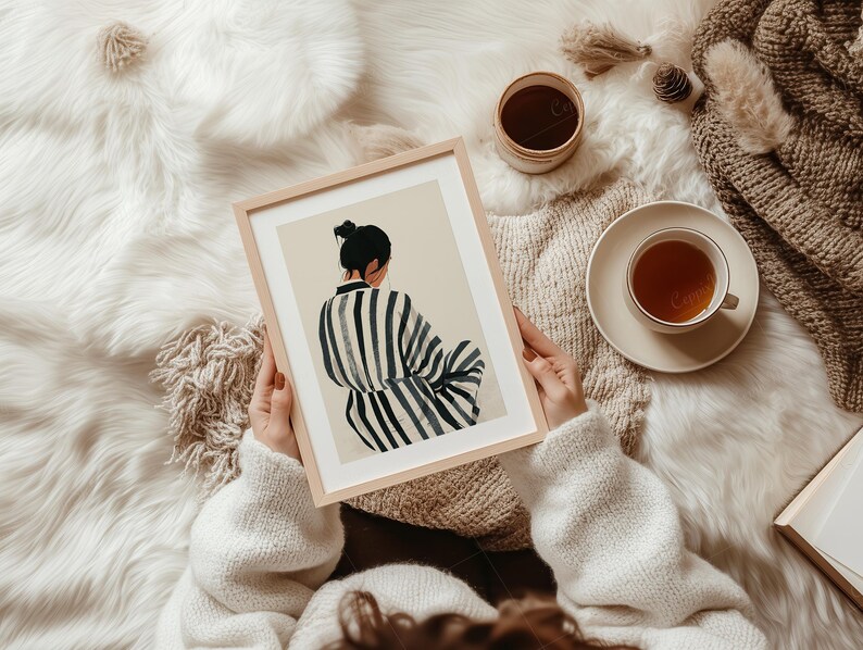 May include: A framed art print featuring a woman in a striped robe, viewed from the back, with a bun hairstyle. The artwork is held by a person wearing a white sweater, with a cup of tea and a decorative blanket in the background.