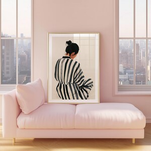 May include: A framed art print featuring a person in a black and white striped garment, seated with their back to the viewer. The artwork is displayed above a pink chaise lounge, set against a backdrop of large windows overlooking a cityscape.