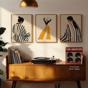 May include: Three framed art prints with abstract figures in neutral tones and gold frames hang above a wooden cabinet. A record player and a vinyl record are on the cabinet. The record cover reads "THE BEATLES A HARD DAY'S NIGHT."