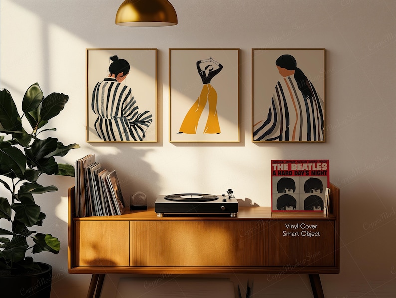 May include: Three framed art prints with abstract figures in neutral tones and gold frames hang above a wooden cabinet. A record player and a vinyl record are on the cabinet. The record cover reads "THE BEATLES A HARD DAY'S NIGHT."