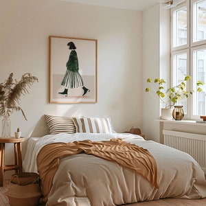 May include: A cozy bedroom scene with a bed covered in neutral-toned bedding and a brown throw blanket. A framed artwork depicting a person in a green top and striped skirt hangs above the bed. A wooden stool and a vase with dried plants add to the room's decor.