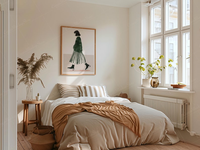 May include: A cozy bedroom scene with a bed covered in neutral-toned bedding and a brown throw blanket. A framed artwork depicting a person in a green top and striped skirt hangs above the bed. A wooden stool and a vase with dried plants add to the room's decor.