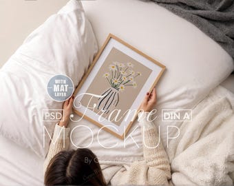 May include: A person holding a wooden frame with a white mat and a print of a bouquet of white daisies in a vase on a beige background. The frame is on a white bed with a white blanket and a gray throw blanket.