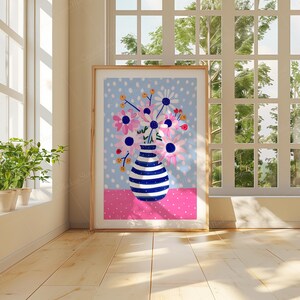 May include: A framed illustration of a blue and white striped vase filled with pink and white flowers on a blue background with white polka dots. The vase is sitting on a pink background with white polka dots.