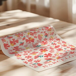 May include: A rolled-up poster featuring a floral pattern with pink and yellow flowers on a cream background. The word "BARCELONA" is printed in red, along with other text. The poster is on a light-colored wooden surface.