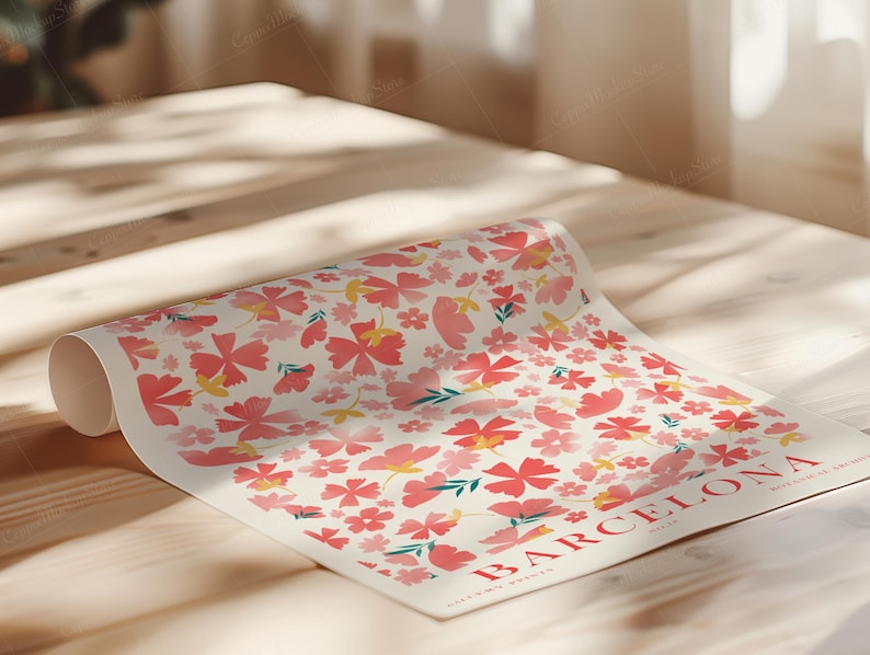 May include: A rolled-up poster featuring a floral pattern with pink and yellow flowers on a cream background. The word "BARCELONA" is printed in red, along with other text. The poster is on a light-colored wooden surface.