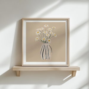 Puede incluir: Una impresi&oacute;n art&iacute;stica enmarcada con margaritas en un jarr&oacute;n. El jarr&oacute;n es blanco con rayas verticales negras. Las margaritas tienen p&eacute;talos blancos y centros amarillos. La obra de arte se exhibe en una estanter&iacute;a de madera clara contra una pared blanca.