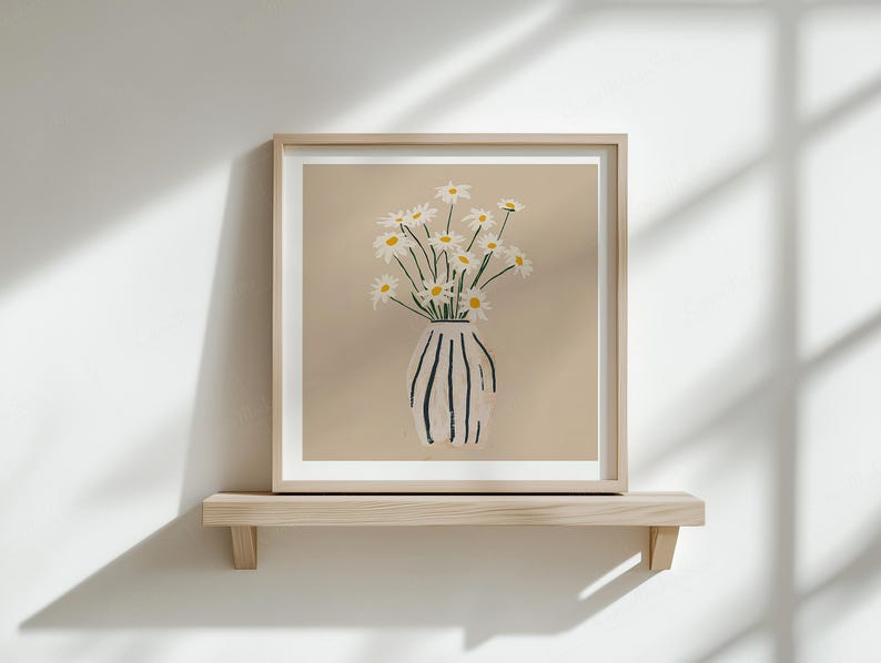 Puede incluir: Una impresi&oacute;n art&iacute;stica enmarcada con margaritas en un jarr&oacute;n. El jarr&oacute;n es blanco con rayas verticales negras. Las margaritas tienen p&eacute;talos blancos y centros amarillos. La obra de arte se exhibe en una estanter&iacute;a de madera clara contra una pared blanca.