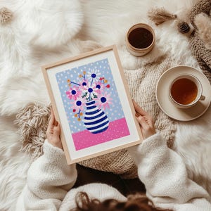 May include: A framed art print featuring a colorful floral arrangement in a striped vase. The artwork has a light blue dotted background, a pink base, and a wooden frame. A cup of tea and a small container are also visible.