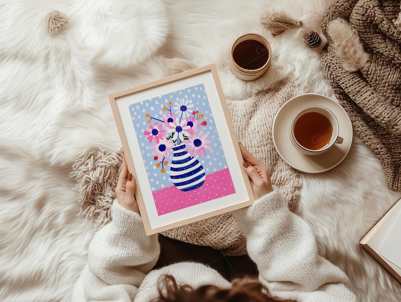 May include: A framed art print featuring a colorful floral arrangement in a striped vase. The artwork has a light blue dotted background, a pink base, and a wooden frame. A cup of tea and a small container are also visible.