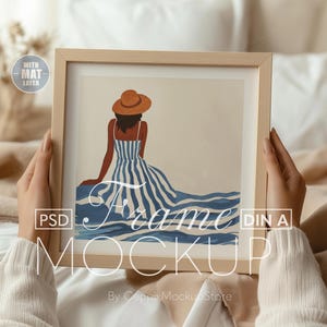 以下が含まれることがあります： 青と白のストライプのドレスと茶色の帽子をかぶった女性が描かれた、額装されたアートプリント。 作品はクリーム色のセーターを着た人が持っています。 フレームは明るい色の木材です。 テキスト「Frame Mockup」が表示されています。