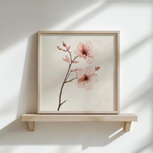 Puede incluir: Una impresi&oacute;n en acuarela enmarcada de flores de cerezo rosas sobre un fondo beige claro. La obra de arte se exhibe en una estanter&iacute;a de madera clara contra una pared blanca. El marco es de color madera clara, y las flores tienen una calidad delicada y transl&uacute;cida.