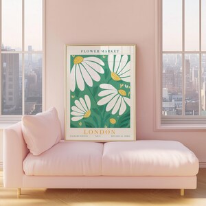 May include: A framed art print featuring a botanical illustration of white daisies with yellow centers on a green background. The print is labeled "LONDON FLOWER MARKET" and is displayed above a pink chaise lounge in a room with large windows.