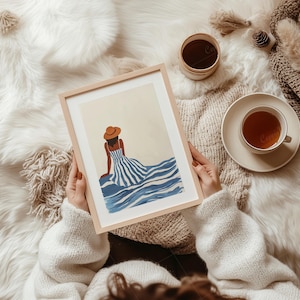 May include: A framed art print featuring a woman in a blue and white striped dress and a brown hat, seated near blue waves. The artwork is held by a person wearing a white sweater. A cup of tea and a small container are also visible.