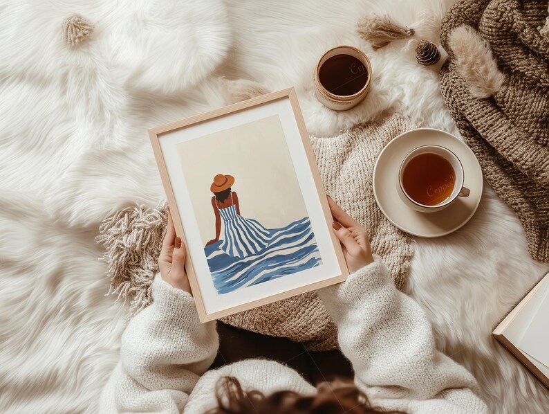 May include: A framed art print featuring a woman in a blue and white striped dress and a brown hat, seated near blue waves. The artwork is held by a person wearing a white sweater. A cup of tea and a small container are also visible.