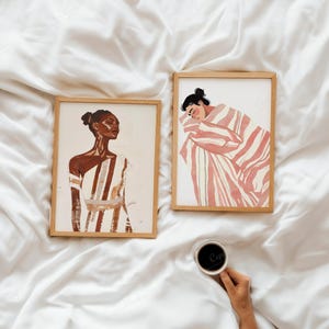 Puede incluir: Dos impresiones art&iacute;sticas enmarcadas con ilustraciones en acuarela. Una representa a una mujer con un vestido a rayas, la otra a una persona envuelta en una manta a rayas. La obra de arte es en tonos marr&oacute;n, blanco y rosa. Una mano sostiene una taza de caf&eacute;.