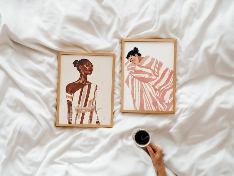 Puede incluir: Dos impresiones art&iacute;sticas enmarcadas con ilustraciones en acuarela. Una representa a una mujer con un vestido a rayas, la otra a una persona envuelta en una manta a rayas. La obra de arte es en tonos marr&oacute;n, blanco y rosa. Una mano sostiene una taza de caf&eacute;.