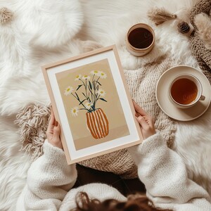 May include: A framed art print featuring a vase of daisies with a light brown background. The vase is orange with vertical stripes. The print is held by a person wearing a white sweater. A cup of tea and a small container are also visible.