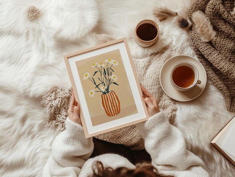 May include: A framed art print featuring a vase of daisies with a light brown background. The vase is orange with vertical stripes. The print is held by a person wearing a white sweater. A cup of tea and a small container are also visible.