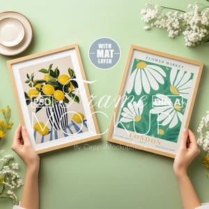 May include: Two framed art prints with botanical themes. One shows lemons in a vase, the other large white daisies on a green background. Both are in light wood frames, held against a pale green backdrop.