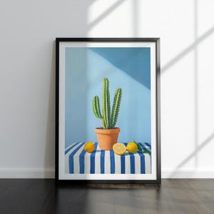 May include: Framed art print showing a cactus in a terracotta pot, set against a light blue backdrop. Lemons are placed on a blue and white striped surface. The artwork is in a black frame.