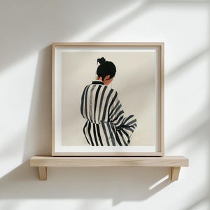 Puede incluir: Impresi&oacute;n art&iacute;stica enmarcada que muestra a una persona con una prenda a rayas, vista por la espalda. La obra de arte se exhibe en una estanter&iacute;a de madera clara contra una pared blanca. Las sombras de la luz natural se proyectan.