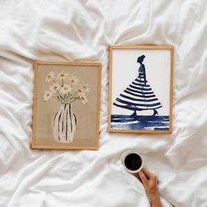 Puede incluir: Dos impresiones art&iacute;sticas enmarcadas sobre una superficie blanca. Una presenta un jarr&oacute;n con margaritas, la otra una figura con un vestido a rayas. Una mano sostiene una taza de caf&eacute;. El arte es de estilo minimalista y acuarela.
