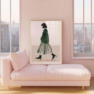 May include: A framed art print featuring a figure in a green sweater and striped skirt, walking. The artwork is displayed on a wall above a pink chaise lounge. The room has large windows with a city view.