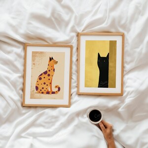 Puede incluir: Dos impresiones art&iacute;sticas enmarcadas con ilustraciones de gatos. Una muestra un gato naranja con manchas moradas sobre un fondo beige. La otra representa un gato negro sobre un fondo dorado. Ambas en marcos de madera.
