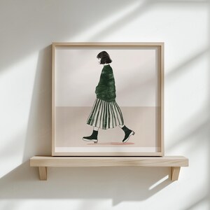 Puede incluir: Una impresi&oacute;n art&iacute;stica enmarcada con una ilustraci&oacute;n minimalista de una persona caminando. La figura lleva un su&eacute;ter verde, una falda a rayas y botas negras. La obra de arte se exhibe en una estanter&iacute;a de madera contra una pared blanca.