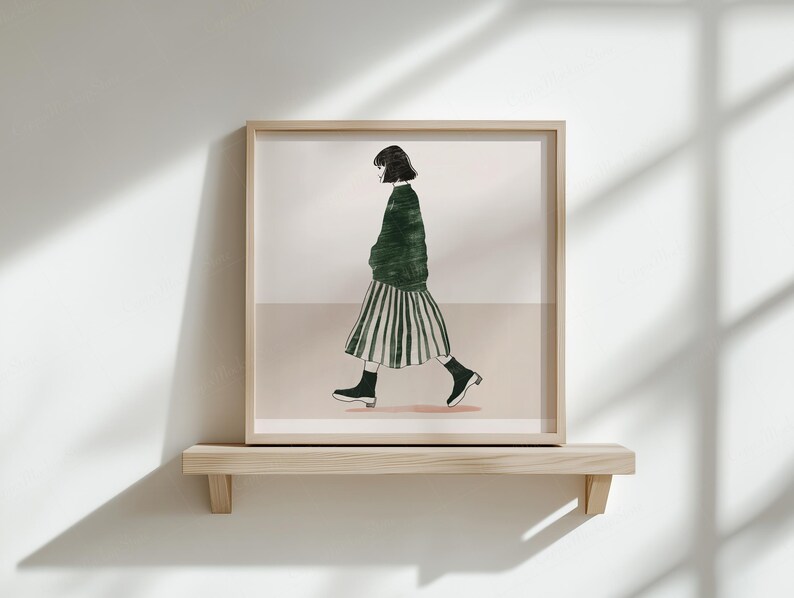 Puede incluir: Una impresi&oacute;n art&iacute;stica enmarcada con una ilustraci&oacute;n minimalista de una persona caminando. La figura lleva un su&eacute;ter verde, una falda a rayas y botas negras. La obra de arte se exhibe en una estanter&iacute;a de madera contra una pared blanca.