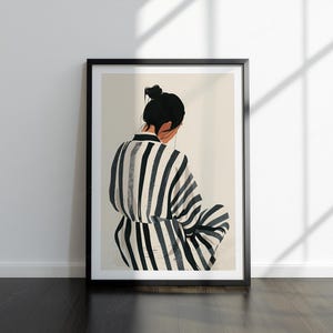 May include: Framed art print showing a person in a striped robe, seen from behind. The figure has dark hair in a bun. The robe has black and white vertical stripes. The artwork is in a black frame, leaning against a white wall.