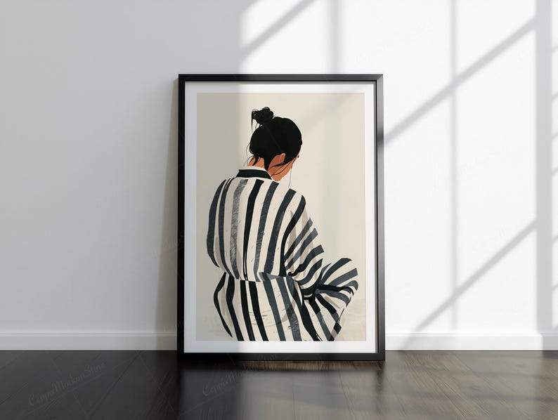 May include: Framed art print showing a person in a striped robe, seen from behind. The figure has dark hair in a bun. The robe has black and white vertical stripes. The artwork is in a black frame, leaning against a white wall.