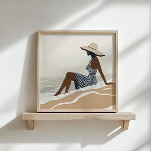 Puede incluir: Una impresi&oacute;n art&iacute;stica enmarcada representa a una persona con un vestido a rayas y un sombrero de ala ancha, sentada en una playa. La obra de arte presenta una paleta de colores neutros con tonos beige, azul y marr&oacute;n. La impresi&oacute;n se exhibe en una estanter&iacute;a de madera contra una pared blanca.