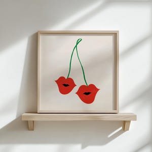 Puede incluir: Una impresi&oacute;n art&iacute;stica enmarcada que presenta dos labios rojos unidos por un tallo verde, parecidos a cerezas. La obra de arte se exhibe en una estanter&iacute;a de madera clara contra una pared blanca, creando una est&eacute;tica minimalista.
