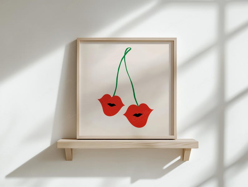 Puede incluir: Una impresi&oacute;n art&iacute;stica enmarcada que presenta dos labios rojos unidos por un tallo verde, parecidos a cerezas. La obra de arte se exhibe en una estanter&iacute;a de madera clara contra una pared blanca, creando una est&eacute;tica minimalista.