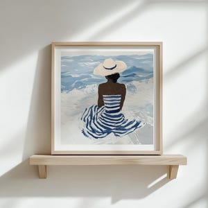 Puede incluir: Una impresi&oacute;n art&iacute;stica enmarcada representa a una persona con un vestido a rayas y un sombrero para el sol, sentada de espaldas al espectador, frente al mar. La obra de arte presenta tonos de azul y blanco, montada en una estanter&iacute;a de madera contra una pared blanca.