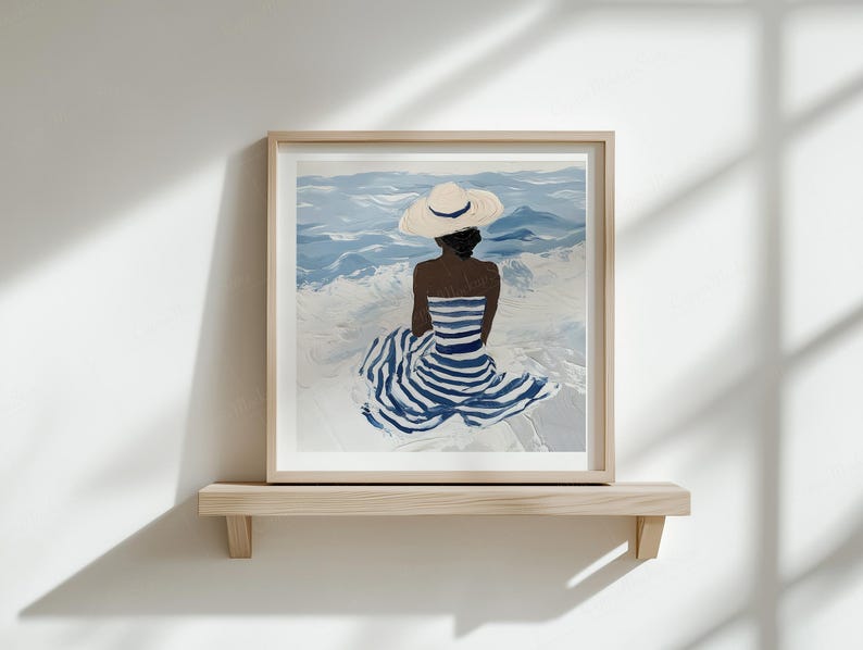 Puede incluir: Una impresi&oacute;n art&iacute;stica enmarcada representa a una persona con un vestido a rayas y un sombrero para el sol, sentada de espaldas al espectador, frente al mar. La obra de arte presenta tonos de azul y blanco, montada en una estanter&iacute;a de madera contra una pared blanca.
