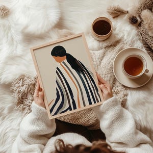 May include: A framed art print featuring an abstract illustration of a person in a striped garment. The artwork has a neutral color palette with black, white, and brown stripes. A cup of tea and a small container are also visible.