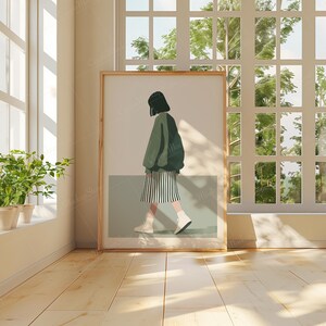 May include: A framed illustration of a person wearing a green jacket and a black and white skirt, walking towards the right side of the image. The person is standing on a light green background. The frame is wooden and has a light brown color.