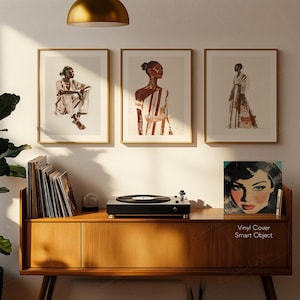 May include: Three framed art prints with gold frames, each featuring a watercolor illustration of a person in fashion. A wooden cabinet holds a record player and vinyl records. A vinyl cover with a woman's face is displayed.