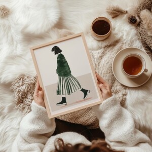 May include: A framed art print featuring a woman in a green jacket and striped skirt. The artwork is held by a person wearing a white sweater, with a cup of tea and a small container nearby. The scene is set on a fluffy white surface.