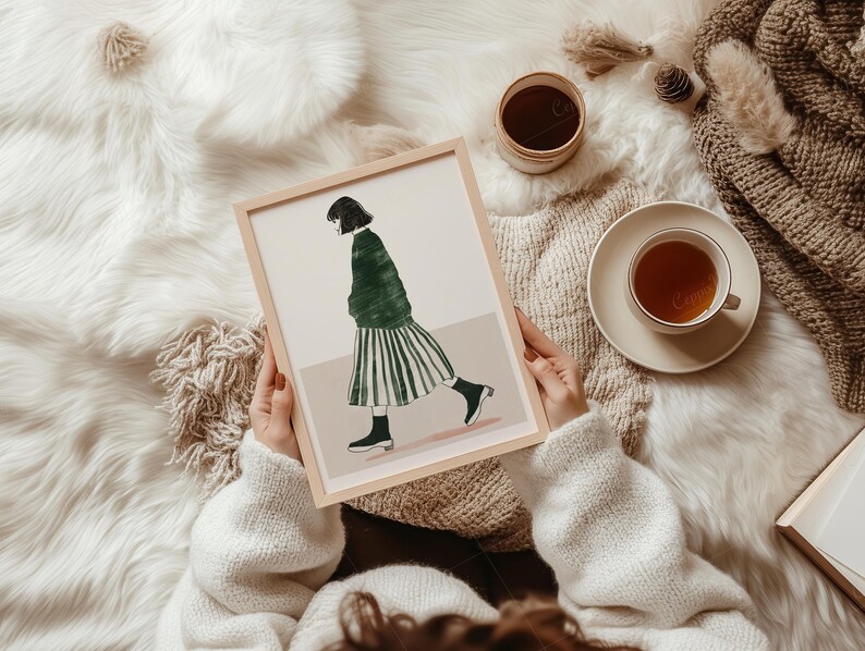 May include: A framed art print featuring a woman in a green jacket and striped skirt. The artwork is held by a person wearing a white sweater, with a cup of tea and a small container nearby. The scene is set on a fluffy white surface.