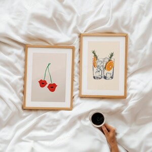 Puede incluir: Dos impresiones art&iacute;sticas enmarcadas con marcos de madera. Una impresi&oacute;n presenta labios rojos como cerezas, la otra muestra dos vasos de c&oacute;ctel con hielo, rodajas de naranja y romero. Una taza de caf&eacute; se sostiene en la mano.