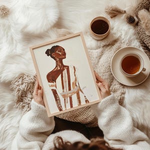 May include: A framed art print featuring a portrait of a person with dark skin, wearing a striped garment. The artwork is held by a person wearing a white sweater. A cup of tea, a small jar, and a blanket are also visible.