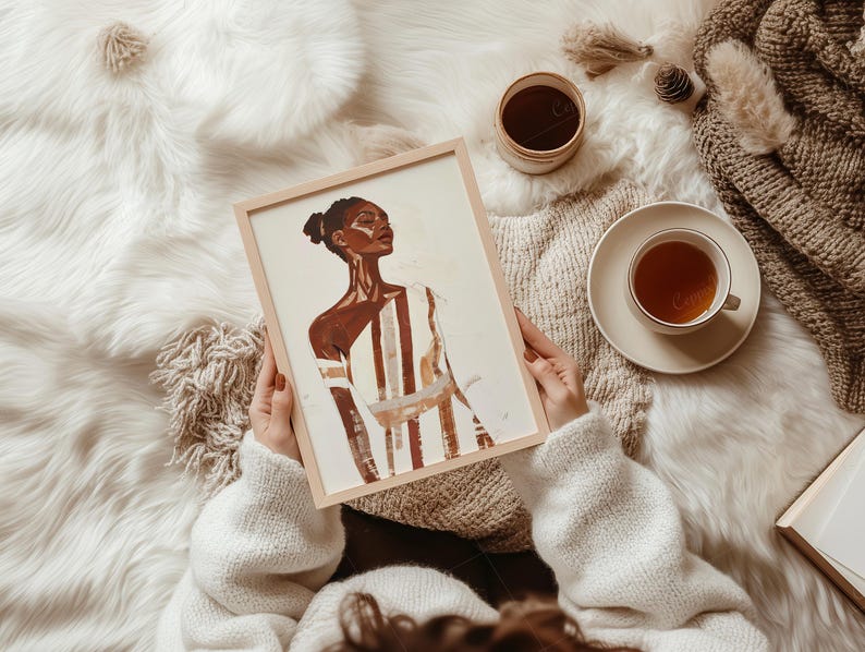 May include: A framed art print featuring a portrait of a person with dark skin, wearing a striped garment. The artwork is held by a person wearing a white sweater. A cup of tea, a small jar, and a blanket are also visible.